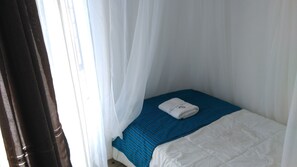 A small, single-bed room with a blue-striped bedspread, white curtains, and a white towel on the bed. - Lago Resort (Kisumu)