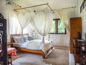 A bedroom with a four-poster bed, a wooden nightstand, a small table with a red tablecloth, and a television on a wooden stand. - Medori Villa (Seminyak)