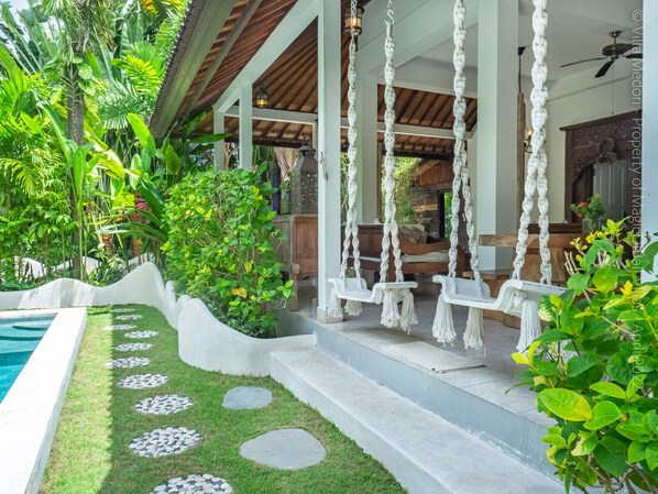 A covered outdoor seating area with white swing chairs and a stone pathway leading to a pool.