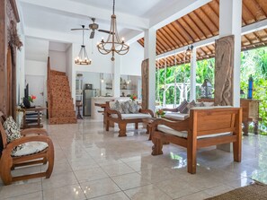 A spacious living area with wooden furniture, a kitchen in the background, and a view of lush greenery through large windows.