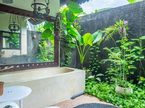 A bathroom with a bathtub, a sink, and a mirror. There is a window with a view of greenery outside. - Medori Villa (Seminyak)