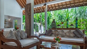 A wooden sofa set with patterned cushions, a wooden coffee table with a bowl and flowers, and a view of lush greenery through a glass door. - Medori Villa (Seminyak)