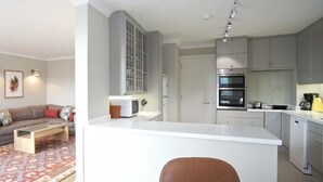 A modern living room with a sofa, coffee table, and lamp, and a kitchen with cabinets and appliances. - A Place Like Home - Chelsea Apartment with River Views (London)