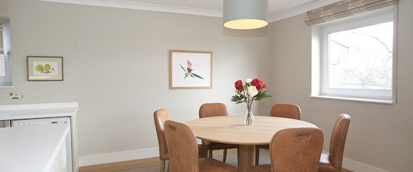 Ein Essbereich mit einem runden Tisch, sechs Stühlen, einer Vase mit Blumen, gerahmten Bildern an der Wand und einem Fenster mit Vorhängen.