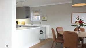 A compact kitchen with a washing machine, a dining area with a table and chairs, and a small seating area with a lamp and books. - A Place Like Home - Chelsea Apartment with River Views (London)