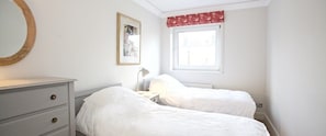 Ein Schlafzimmer mit zwei Betten, einem Schminktisch, einem Nachttisch mit Lampe, einem Fenster mit rotem Vorhang und einem gerahmten Bild an der Wand.