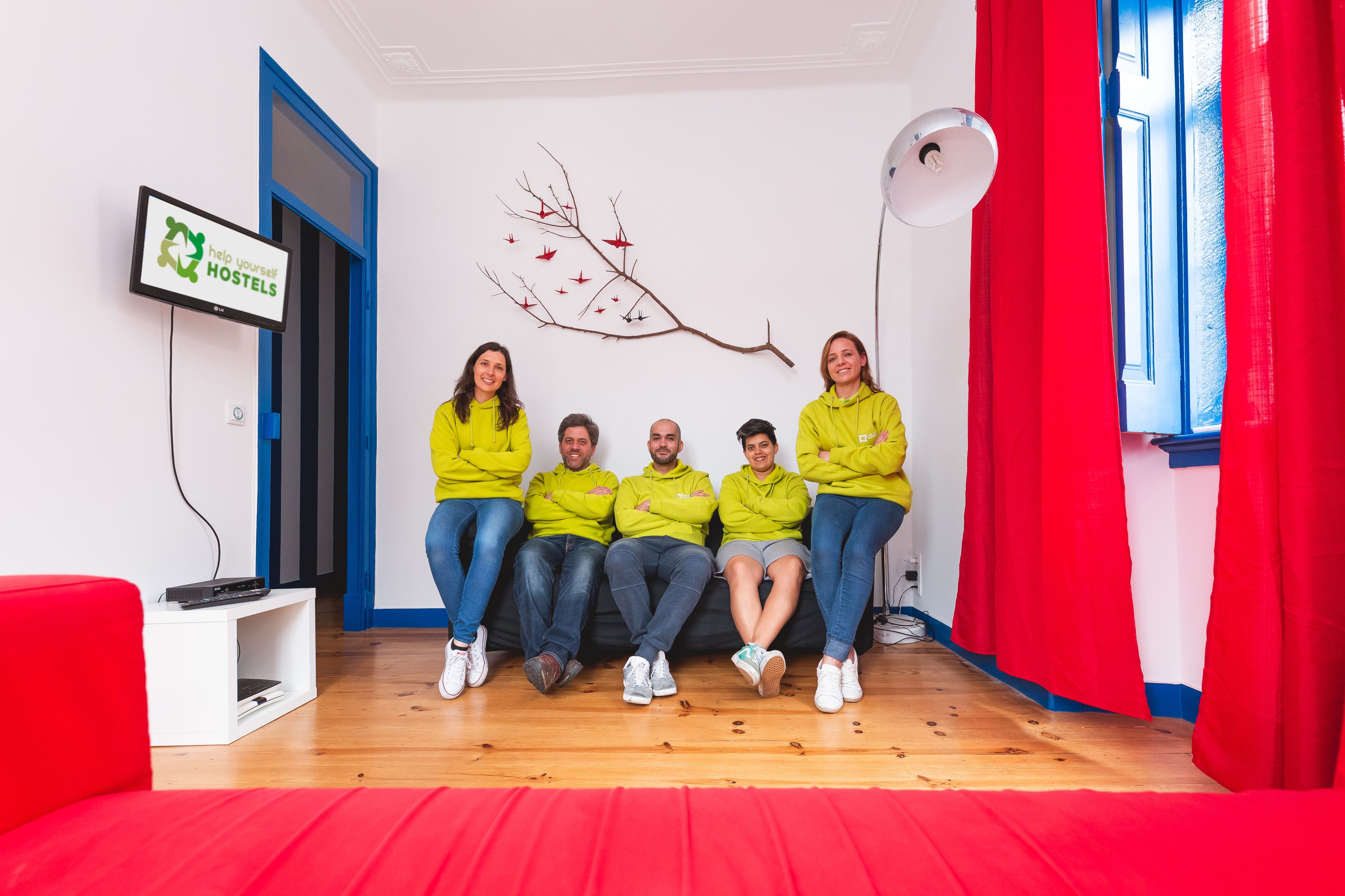 A group of people sitting on a couch in a room with a TV displaying 'help yourself HOSTELS' and a branch decoration on the wall.
