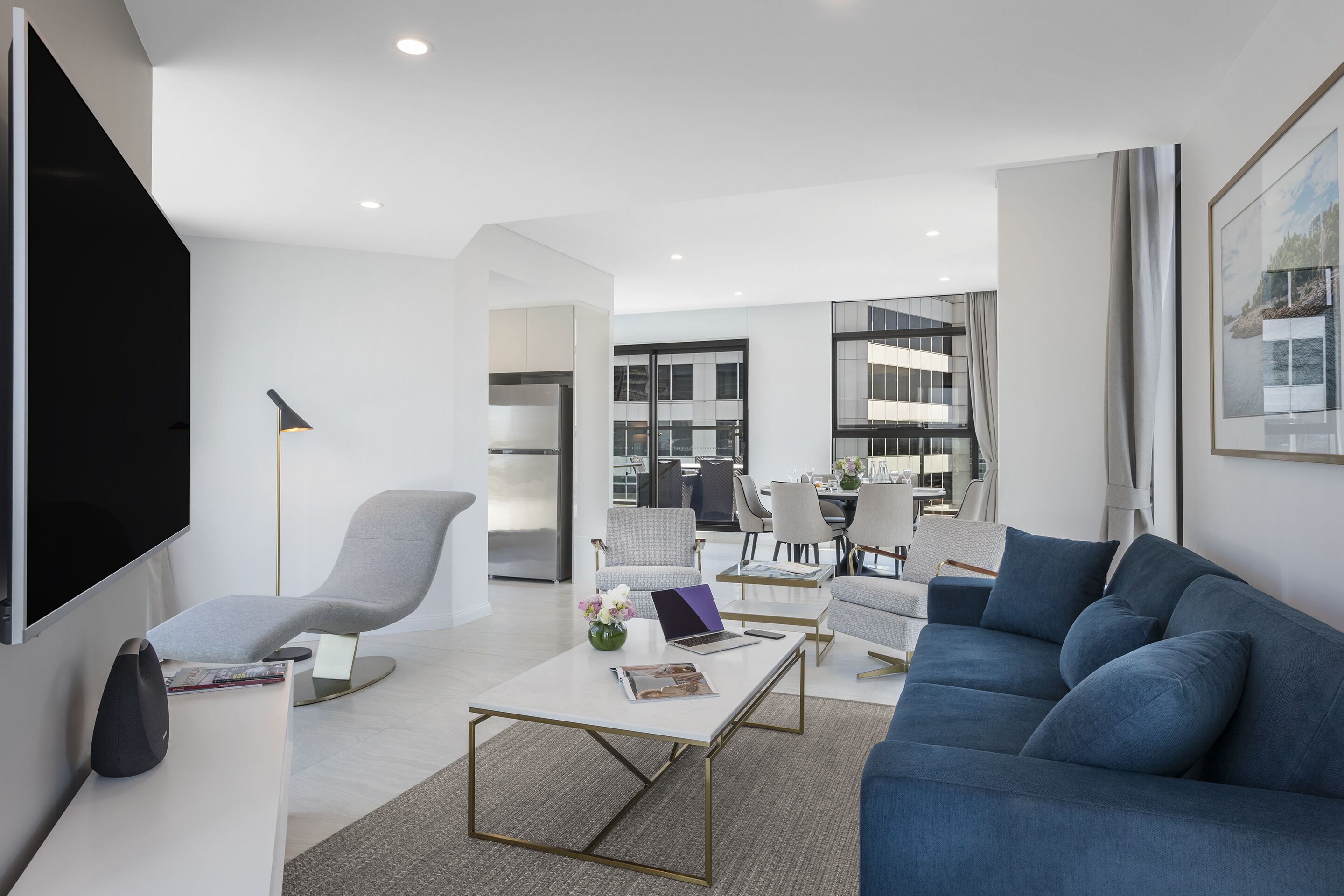 A modern living room with a blue sofa, a grey armchair, and a coffee table. There is a flat-screen TV mounted on the wall and a laptop on the coffee table.