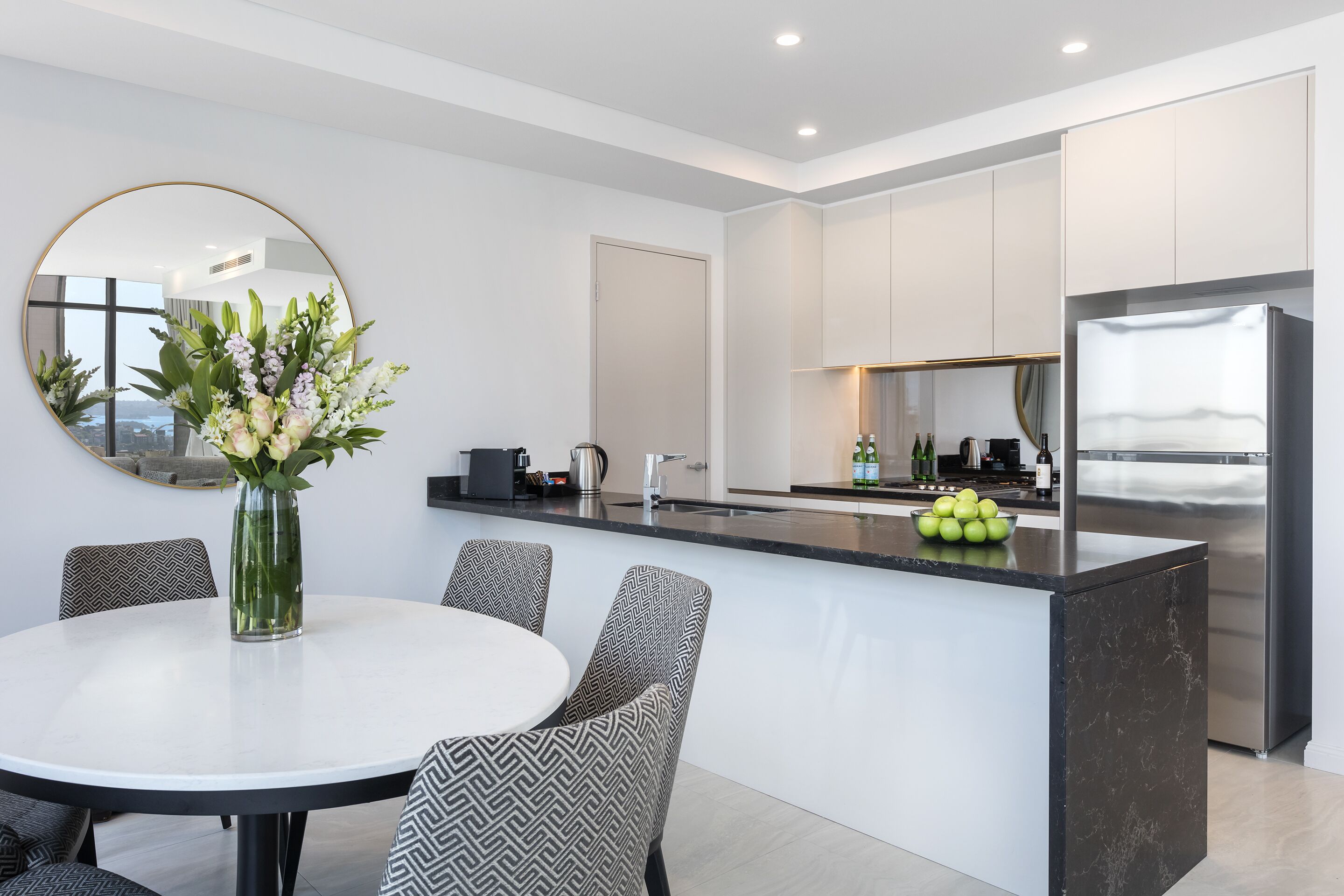 A modern kitchen with a central island, a round dining table with patterned chairs, and a large mirror on the wall.