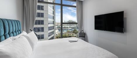 A hotel room with a large bed, a flat-screen TV, a view of the city, and a window with curtains.