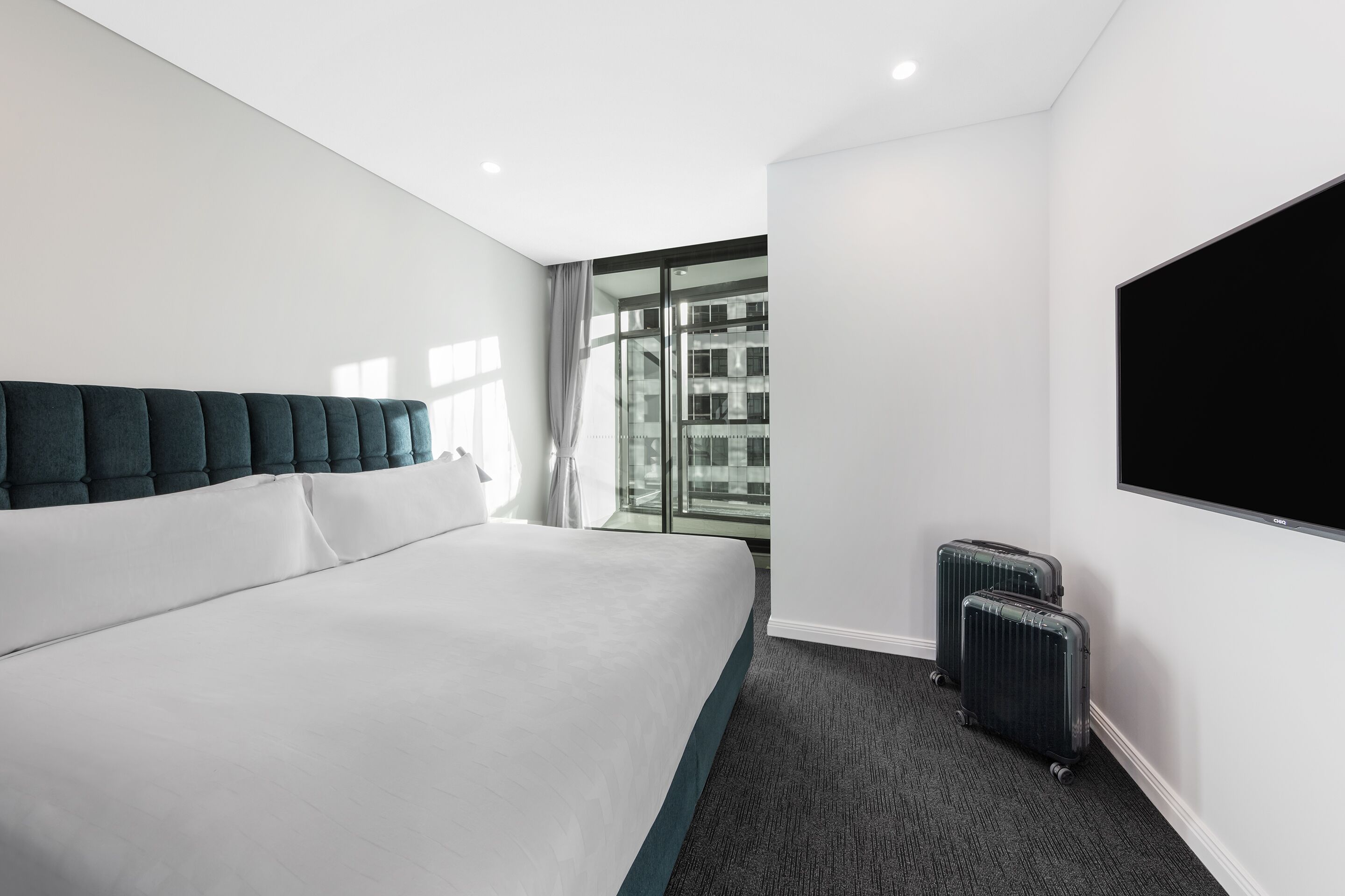 Habitación de hotel con una cama grande, un televisor, una ventana con vistas a edificios y una maleta.