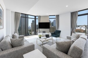 Uma sala de estar moderna com sofás cinzentos, uma mesa de centro, uma TV de tela plana e amplas janelas com vista para a cidade.