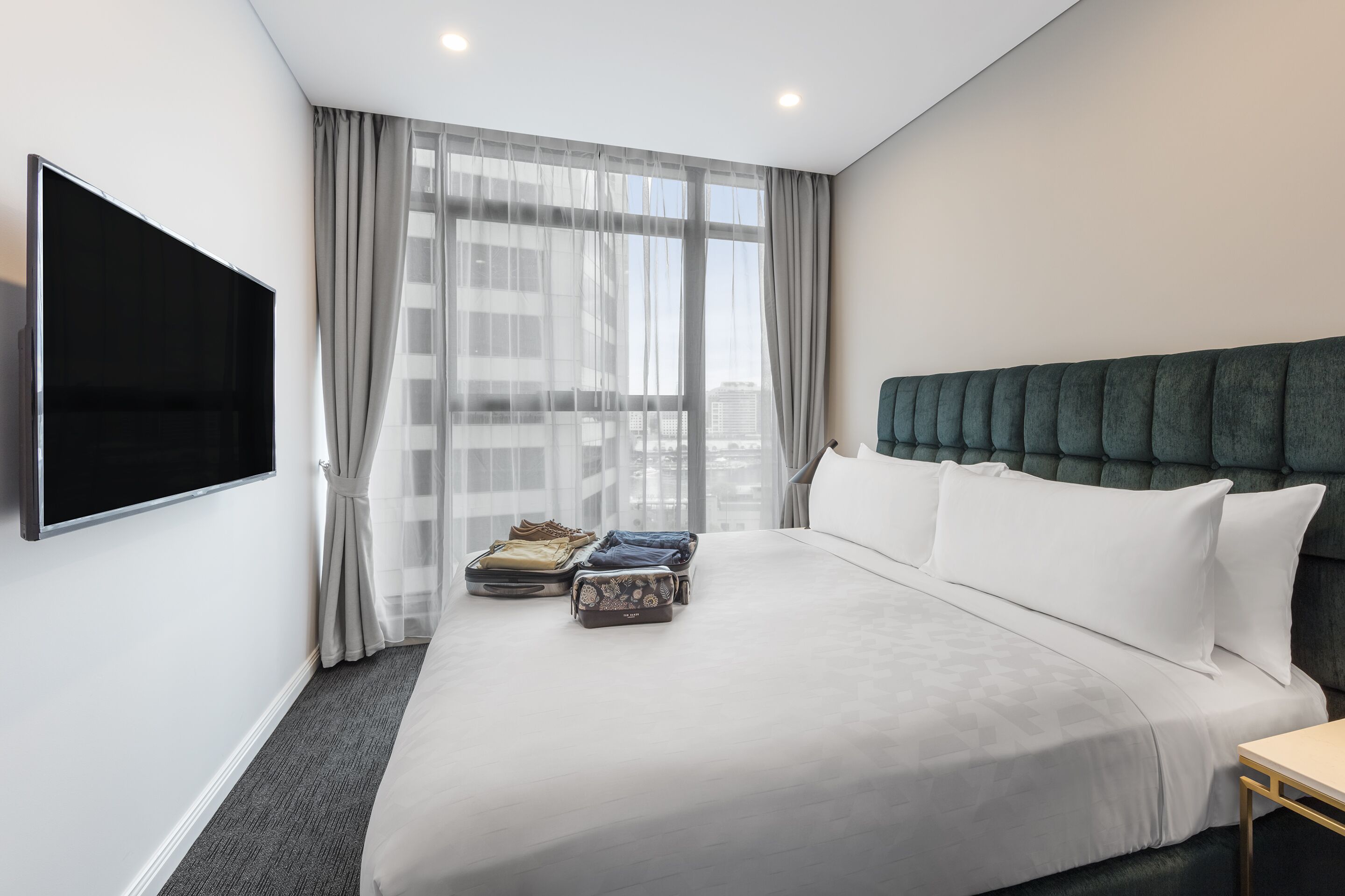 A modern hotel room with a large bed, a flat-screen TV mounted on the wall, and a view of the city through sheer curtains.