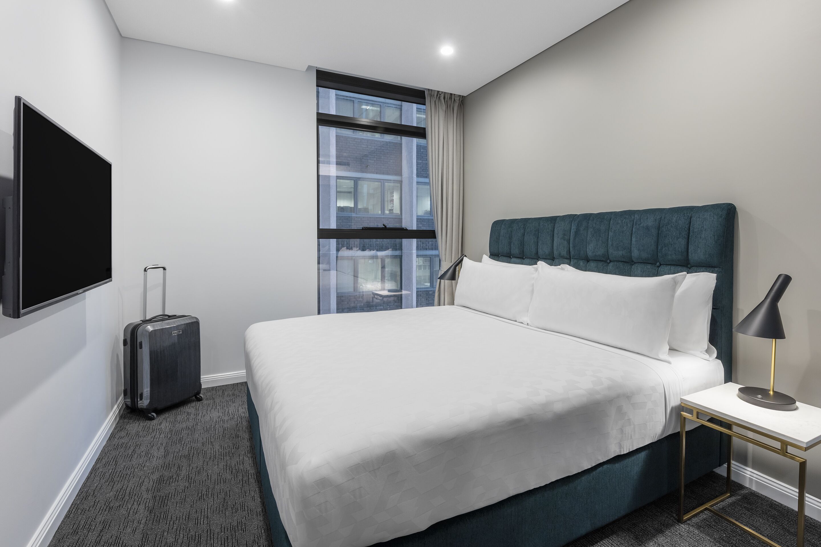 Hotelkamer met een groot bed, een aan de muur bevestigde televisie, een koffer en een raam met uitzicht op gebouwen.