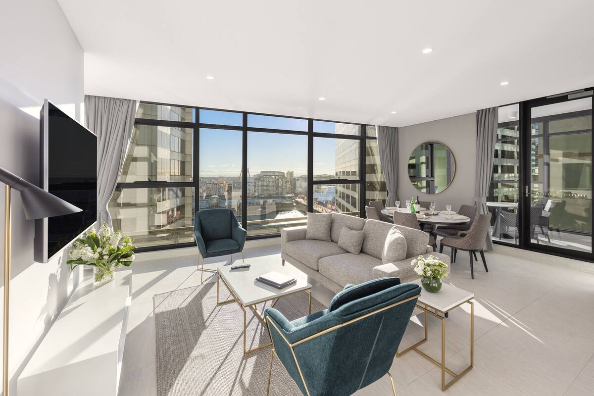 Una sala de estar moderna con un sofá, sillones, una mesa de centro y una televisión. Amplios ventanales ofrecen vistas a la ciudad.