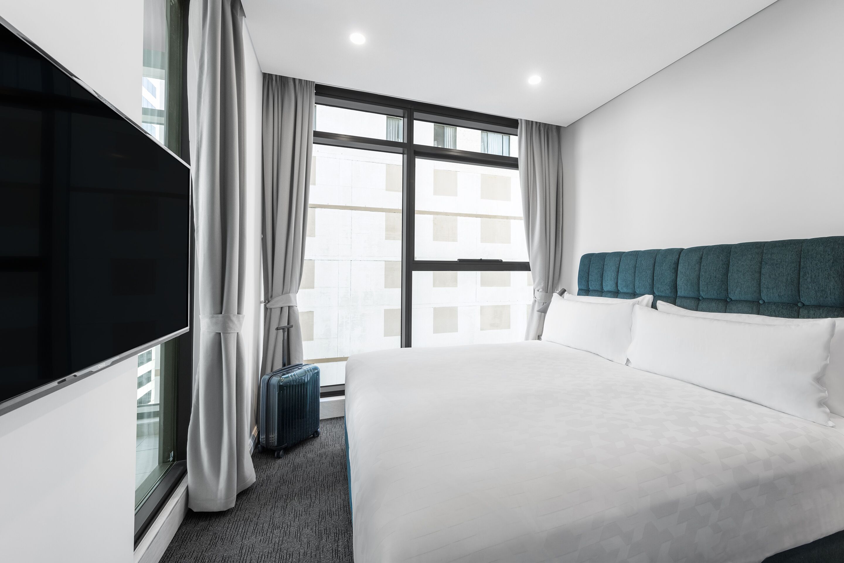 A hotel room with a large bed, a television, a window with curtains, and a suitcase.