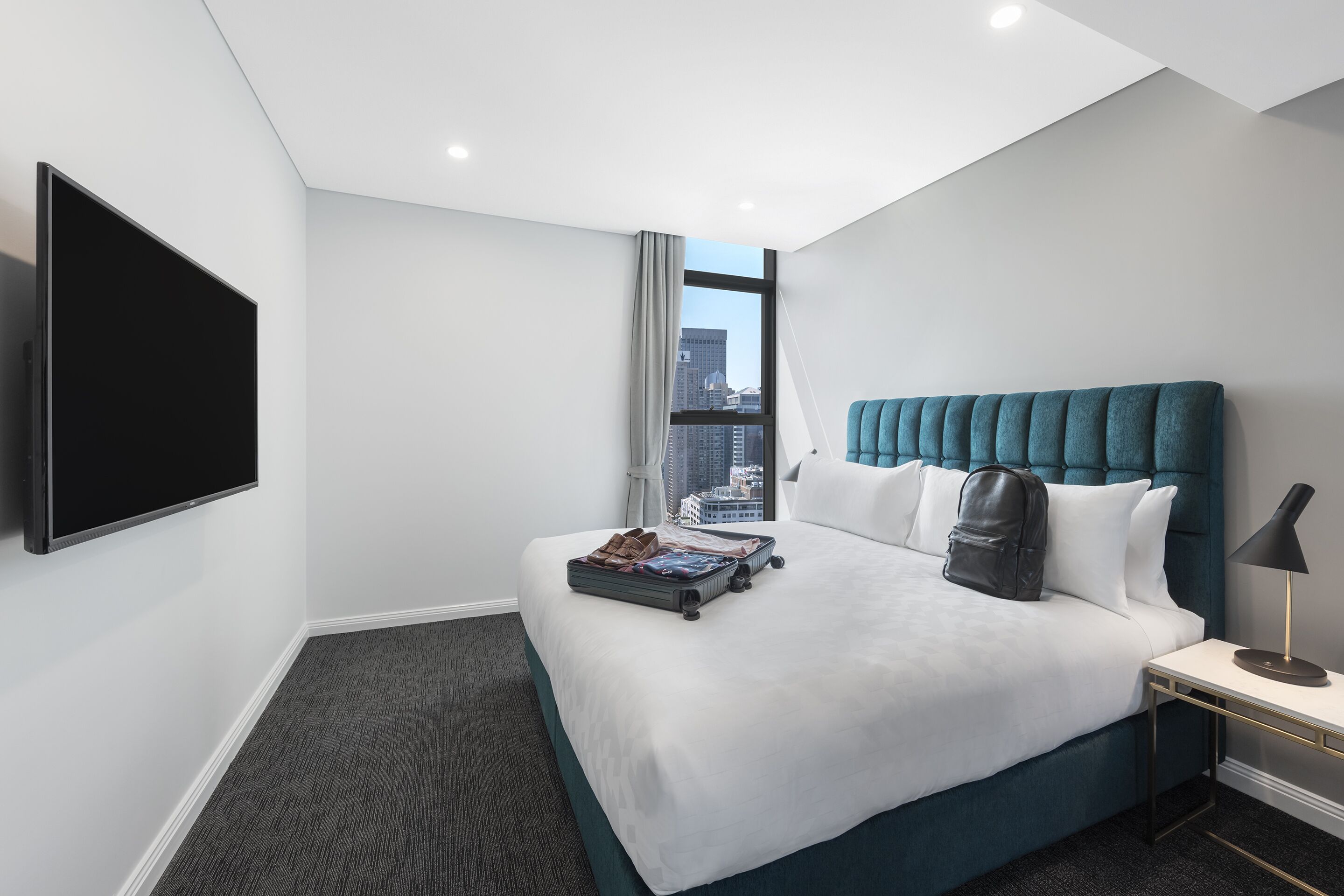 A hotel room with a large bed, a flat-screen TV, a window with a city view, and a bedside table with a lamp.