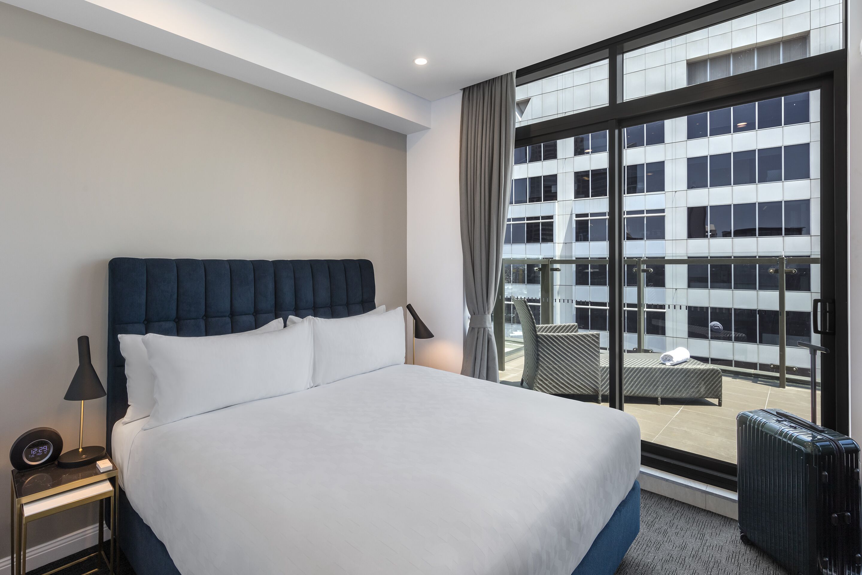 A hotel room with a large bed, a nightstand, a lamp, and a view of a building through a large window.