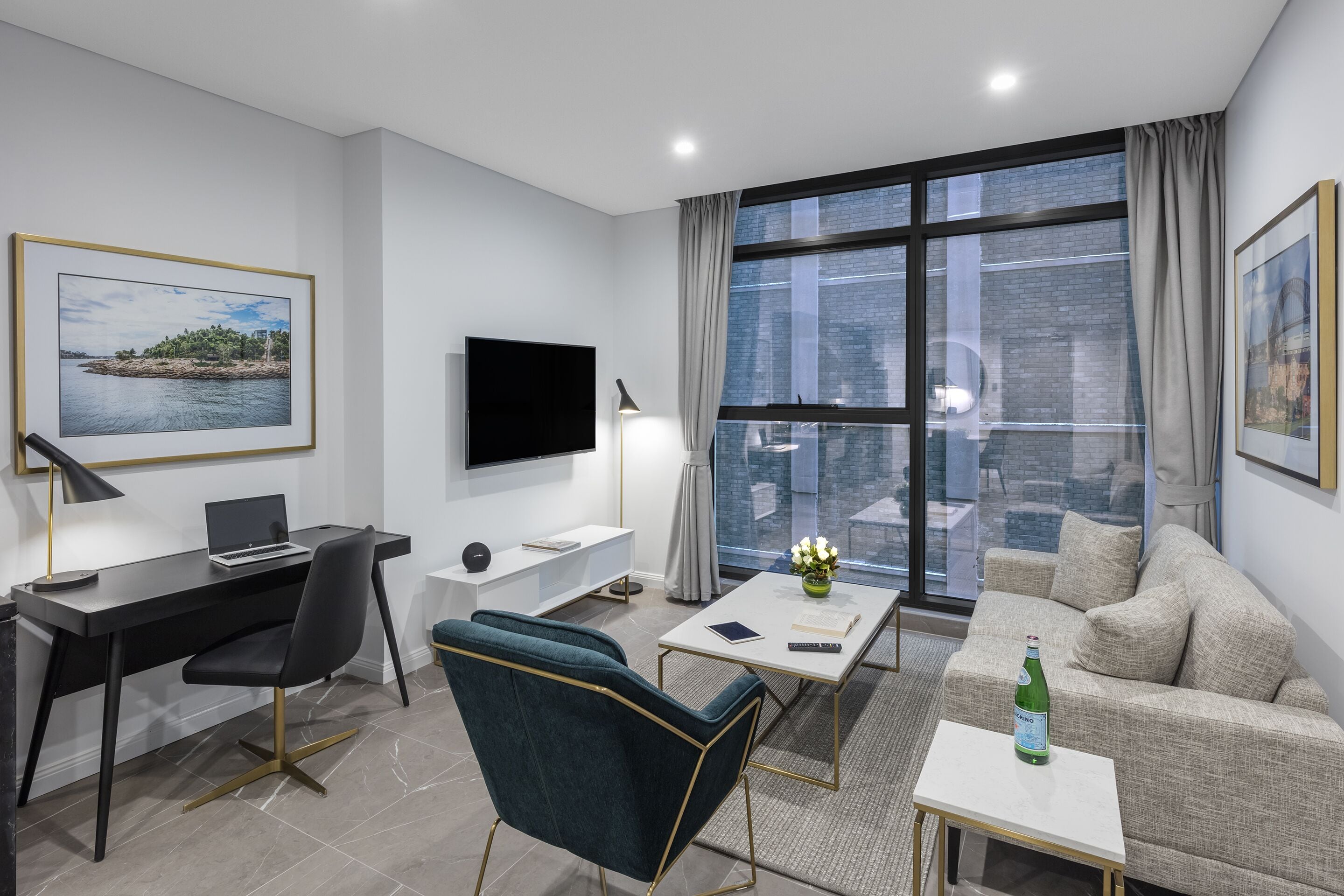 Une chambre d’hôtel moderne dotée d’une grande fenêtre, d’un téléviseur à écran plat, d’un bureau avec un ordinateur portable, d’un canapé confortable et d’une petite table sur laquelle se trouve une bouteille.