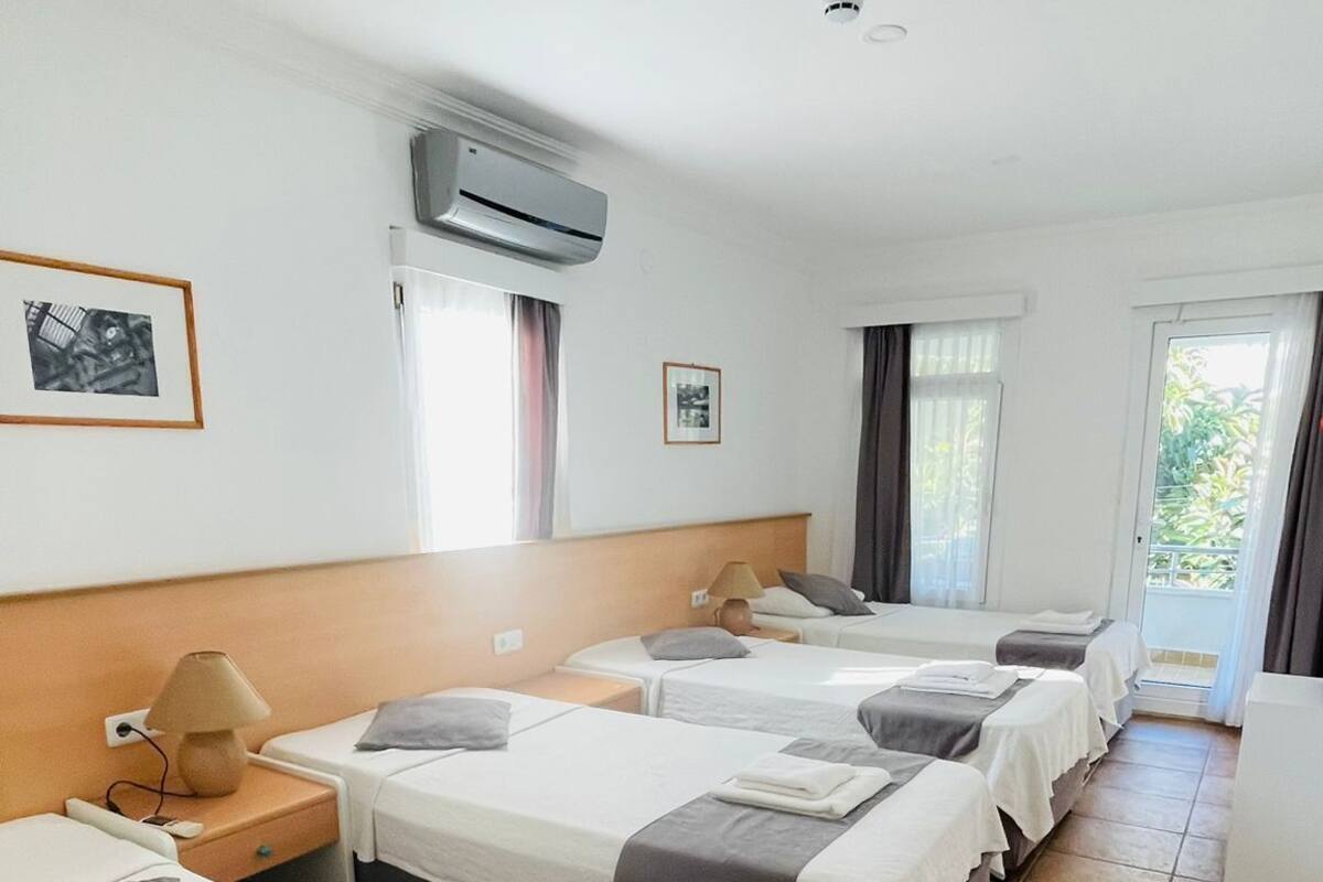 Habitación de hotel con cuatro camas dispuestas en fila, un equipo de aire acondicionado sobre las camas, una ventana con cortinas y una puerta que da al exterior.