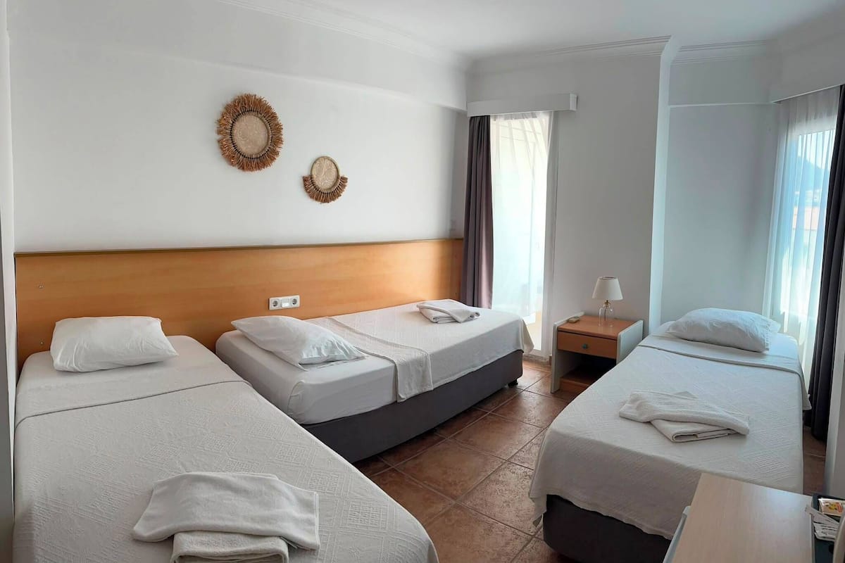 Habitación de hotel con tres camas, cabecera de madera, mesita de noche con lámpara y un reloj de pared.