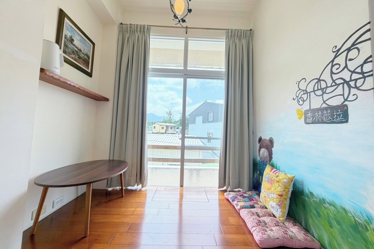 Una habitación con piso de madera, una mesa redonda, un sofá con cojines de colores, un estante en la pared con un cuadro enmarcado y un ventanal con cortinas.