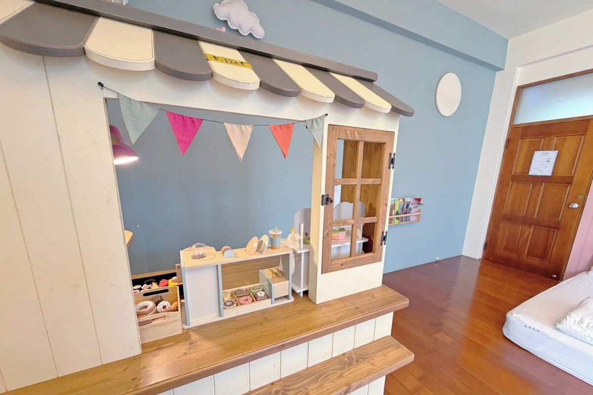 Une pièce avec un plancher en bois, un lit blanc, un petit comptoir en bois avec une vitrine, et une porte avec un panneau en bois.