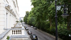 Une vue de rue avec une rangée de bâtiments blancs, un trottoir bordé d’arbres et des voitures garées.