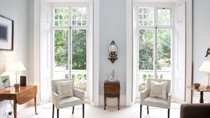 Une pièce avec deux chaises blanches, une petite table et une cheminée. Il y a deux fenêtres à cadres blancs, donnant sur un paysage verdoyant.
