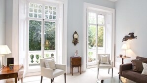 Un salon avec un canapé marron, deux fauteuils blancs, une table basse en verre et une table d’appoint avec une lampe. Il y a deux fenêtres avec des rideaux blancs, une horloge murale et un tableau encadré.