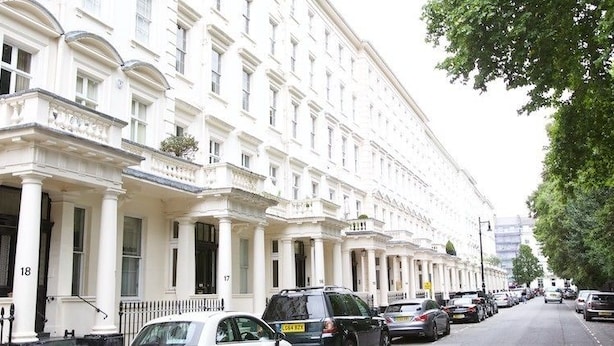 Une rangée de bâtiments blancs au style classique, tous dotés de la même architecture, des voitures garées le long de la rue et un trottoir bordé d’arbres.