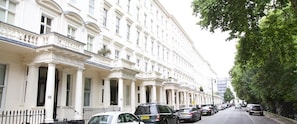 A row of white, classic buildings with uniform architecture, parked cars along the street, and a tree-lined sidewalk.