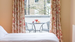 Ein Schlafzimmer mit einem Bett, einem Fenster mit blumigen Vorhängen und einem kleinen Tisch mit einer roten Tasse und einem Teekessel.