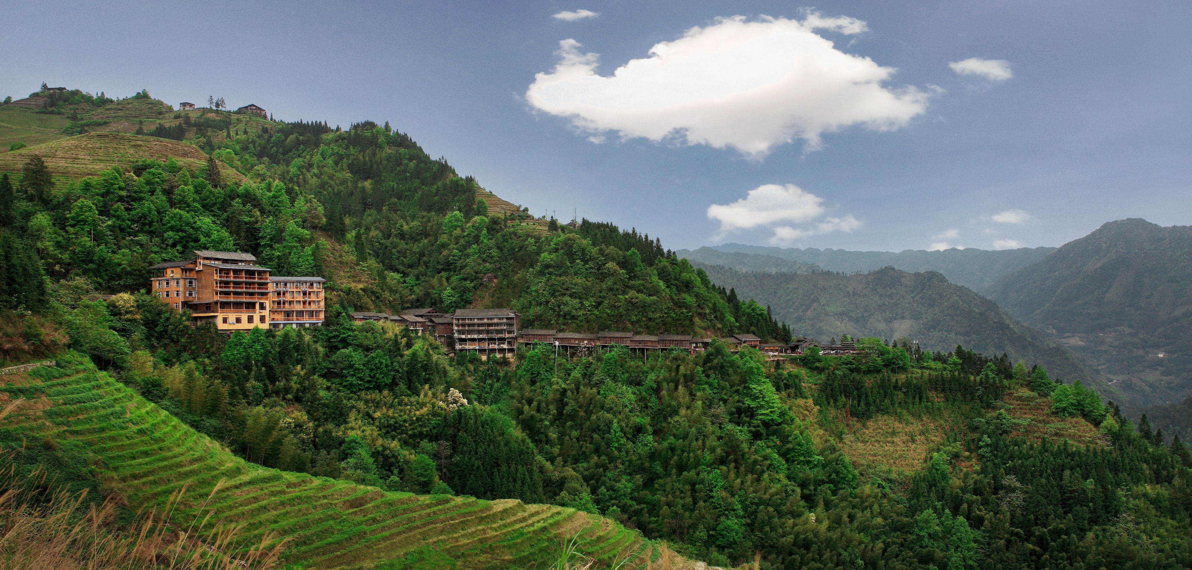 A hillside hotel with multiple buildings, terraced fields, and mountainous terrain in the background.