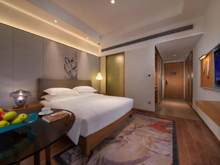 A hotel room with a large bed, a bedside table with a lamp, a fruit bowl, and a painting on the wall.