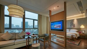 Una habitación de hotel moderna con un televisor grande, asientos cómodos, vistas a la ciudad y una cama con una mesita de noche.