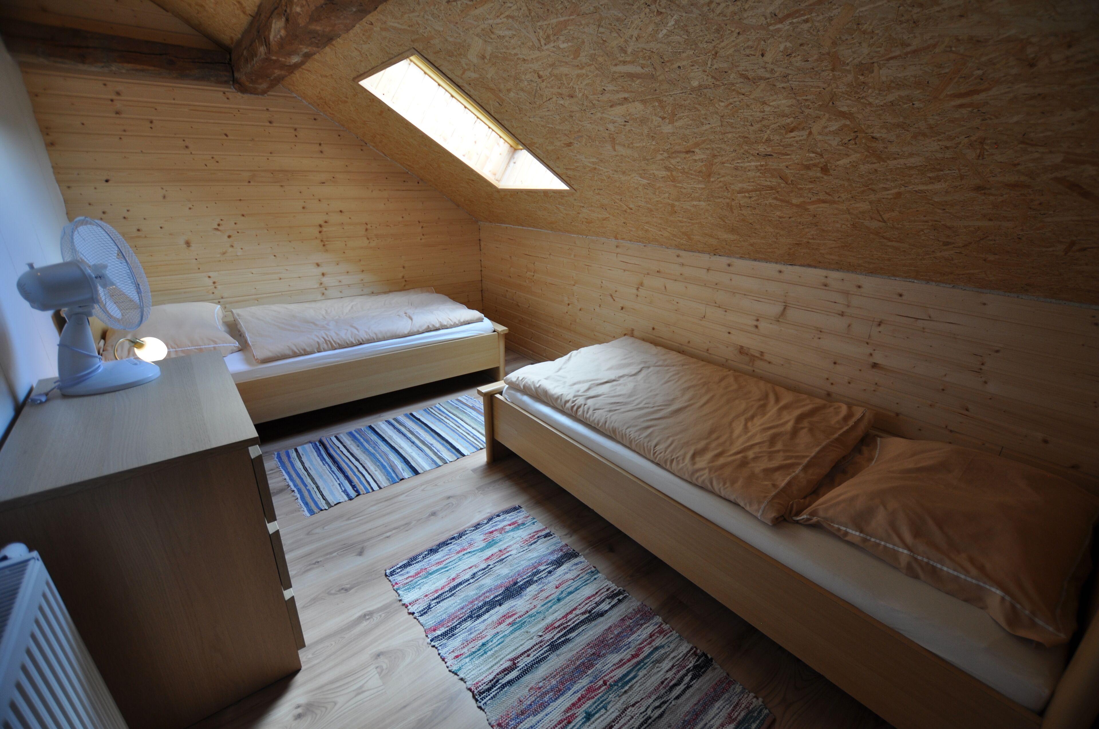 Um quarto pequeno e acolhedor com paredes de madeira e teto inclinado, com duas camas, uma escrivaninha e um ventilador, e uma janela que proporciona luz natural.