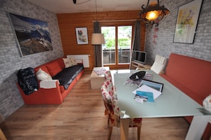 Un salon chaleureux avec un canapé rouge, une table avec des livres et une vue à travers une porte vitrée.