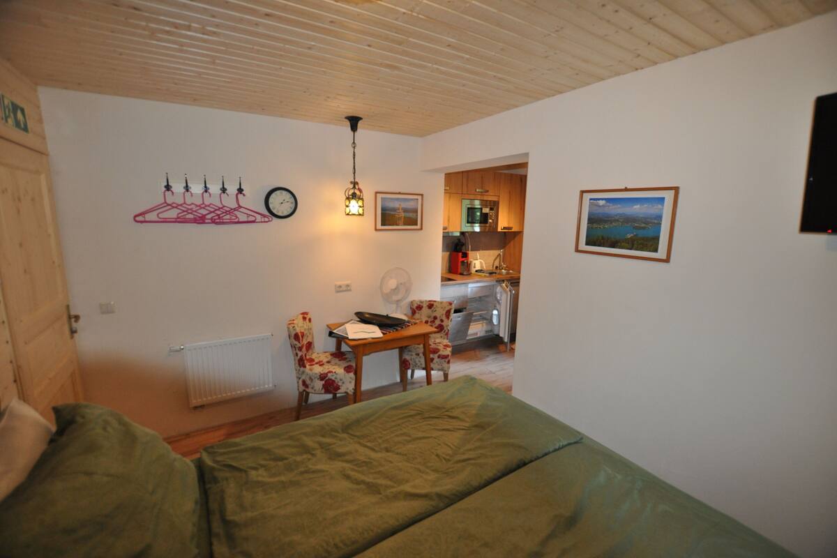 Ein Zimmer mit Bett, Schreibtisch, Stuhl, Heizkörper, einer Uhr und einem durch eine offene Tür einsehbarer Küchenbereich.
