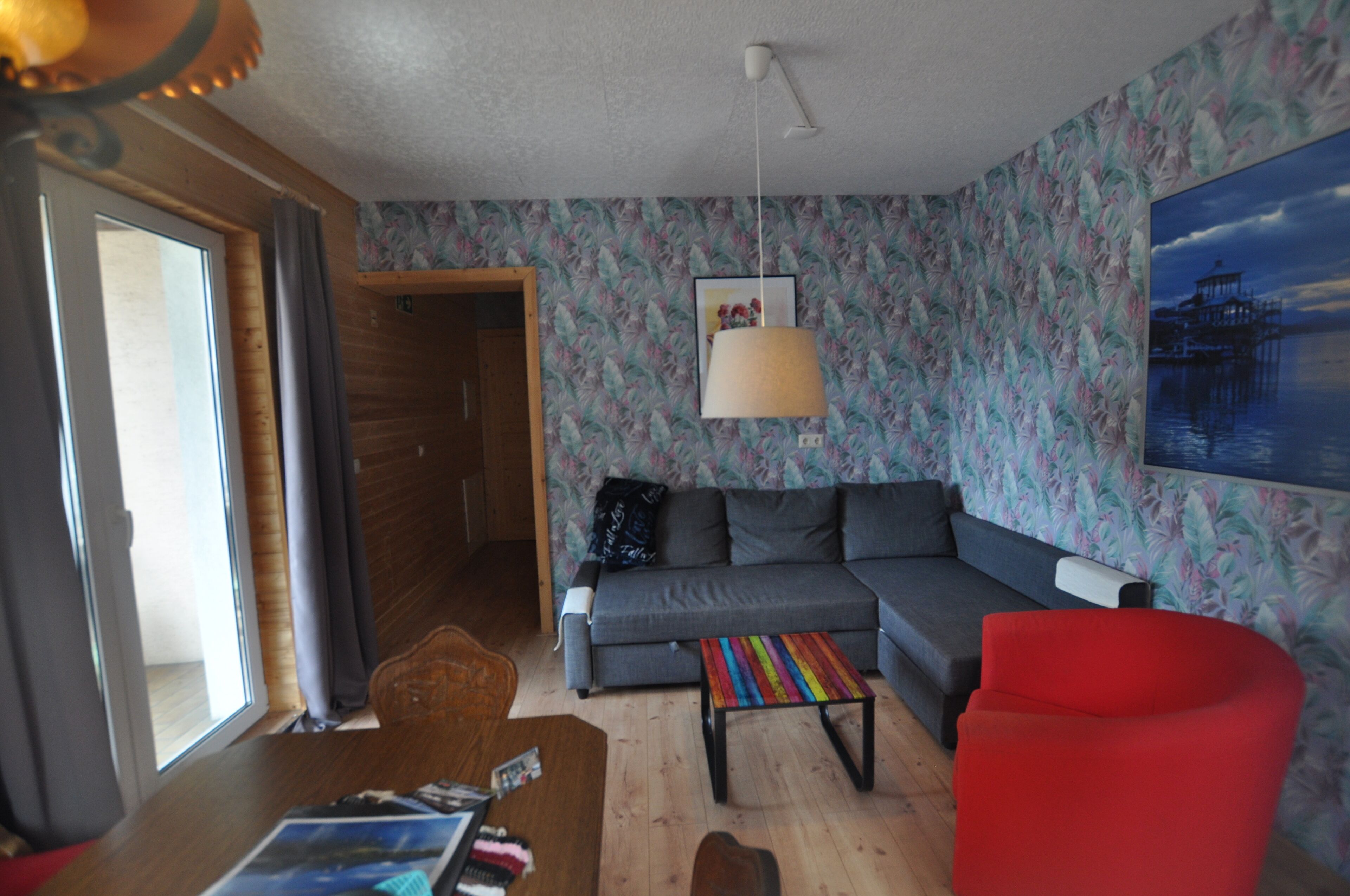 A living room with a grey sofa, a colorful striped coffee table, a red armchair, a wooden door, and a wall with a floral wallpaper.