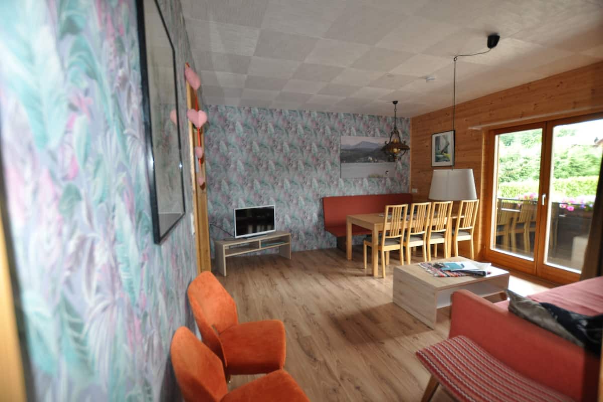 Ein Zimmer mit Holzboden, einem Fernseher auf einem Standfuß, einem Esstisch mit Stühlen, einer Couch und einem Fenster mit Blick auf Grünflächen.