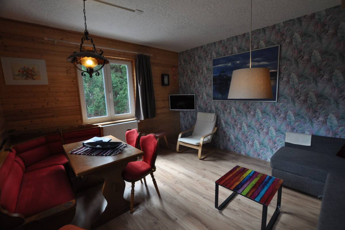 Ein gemütliches Zimmer mit einem Holzesstisch, roten Stühlen, einem grauen Sofa und einem farbenfrohen gestreiften Hocker.