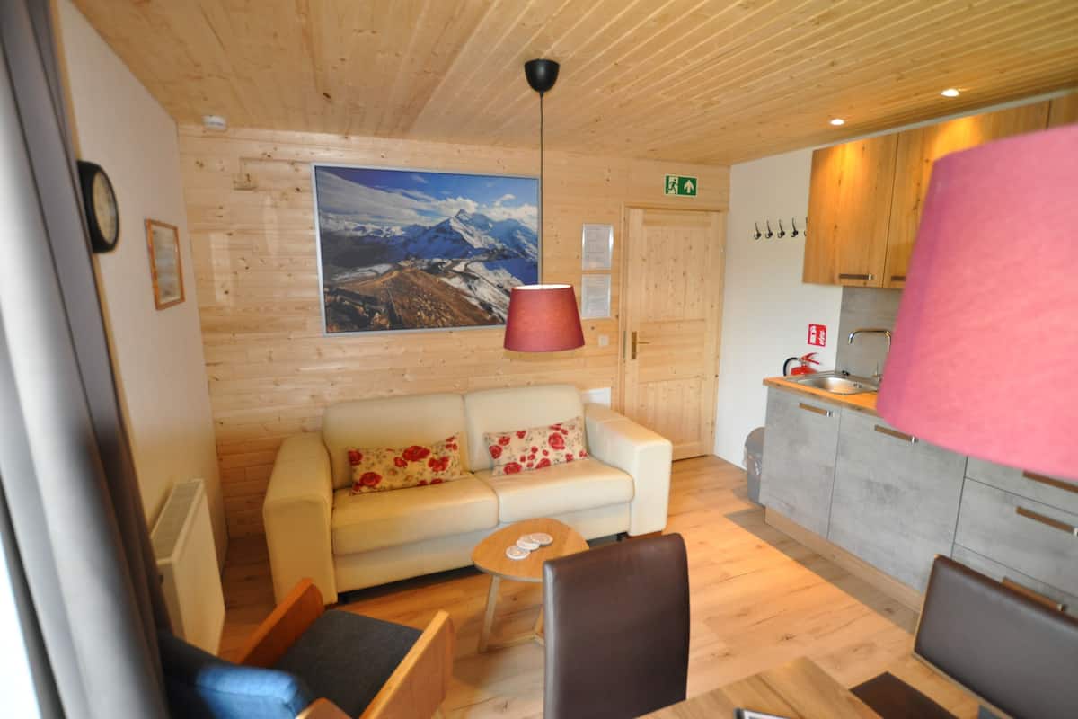 Ein gemütliches Zimmer mit Holzbalkendecke, einer Couch mit floralen Kissen, einem kleinen Tisch und einer Küchenzeile mit Schränken und Spüle.