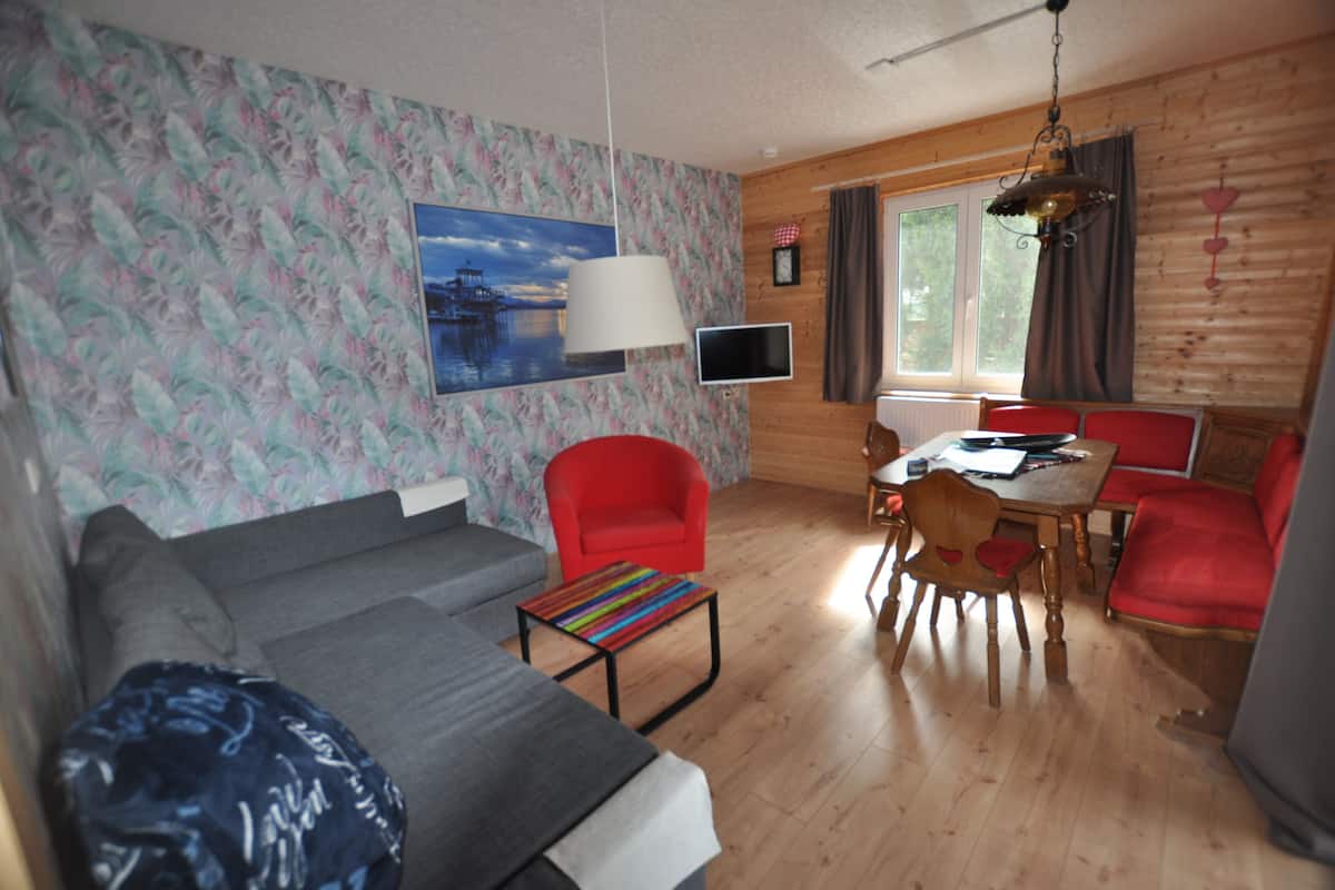 Ein gemütliches Wohnzimmer mit einer Couch, einem roten Sessel, einem Holzesstisch mit Stühlen, einem Fernseher und einem Bild an der Wand.
