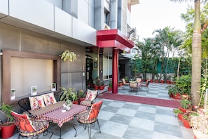 Un espace extérieur aménagé avec des tables recouvertes de nappes à carreaux et des chaises en métal, entouré de plantes en pot et surmonté d’un auvent rouge.