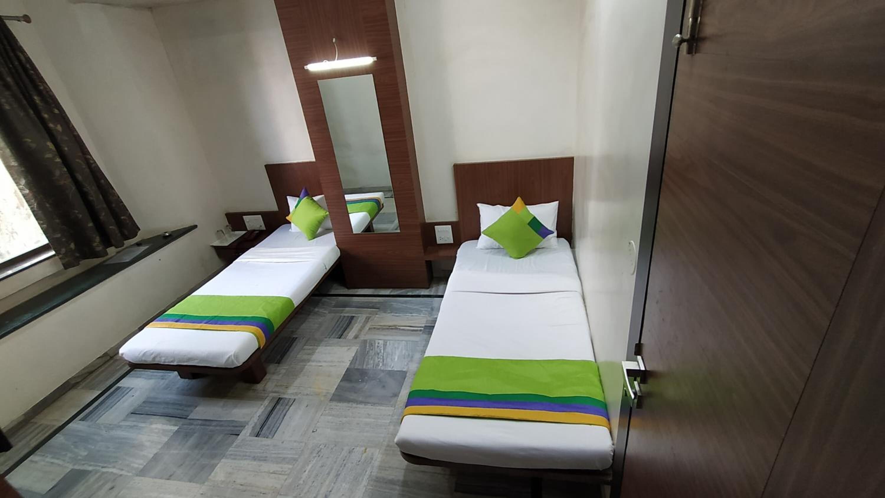 A hotel room with two single beds, wooden headboards, a mirror, a window with curtains, and a tiled floor.