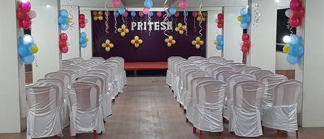A room with rows of chairs covered in white cloth, a stage with a purple backdrop, and colorful balloons.