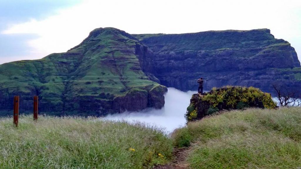 A natural landscape with a grassy foreground, a rocky cliff, and a misty waterfall.