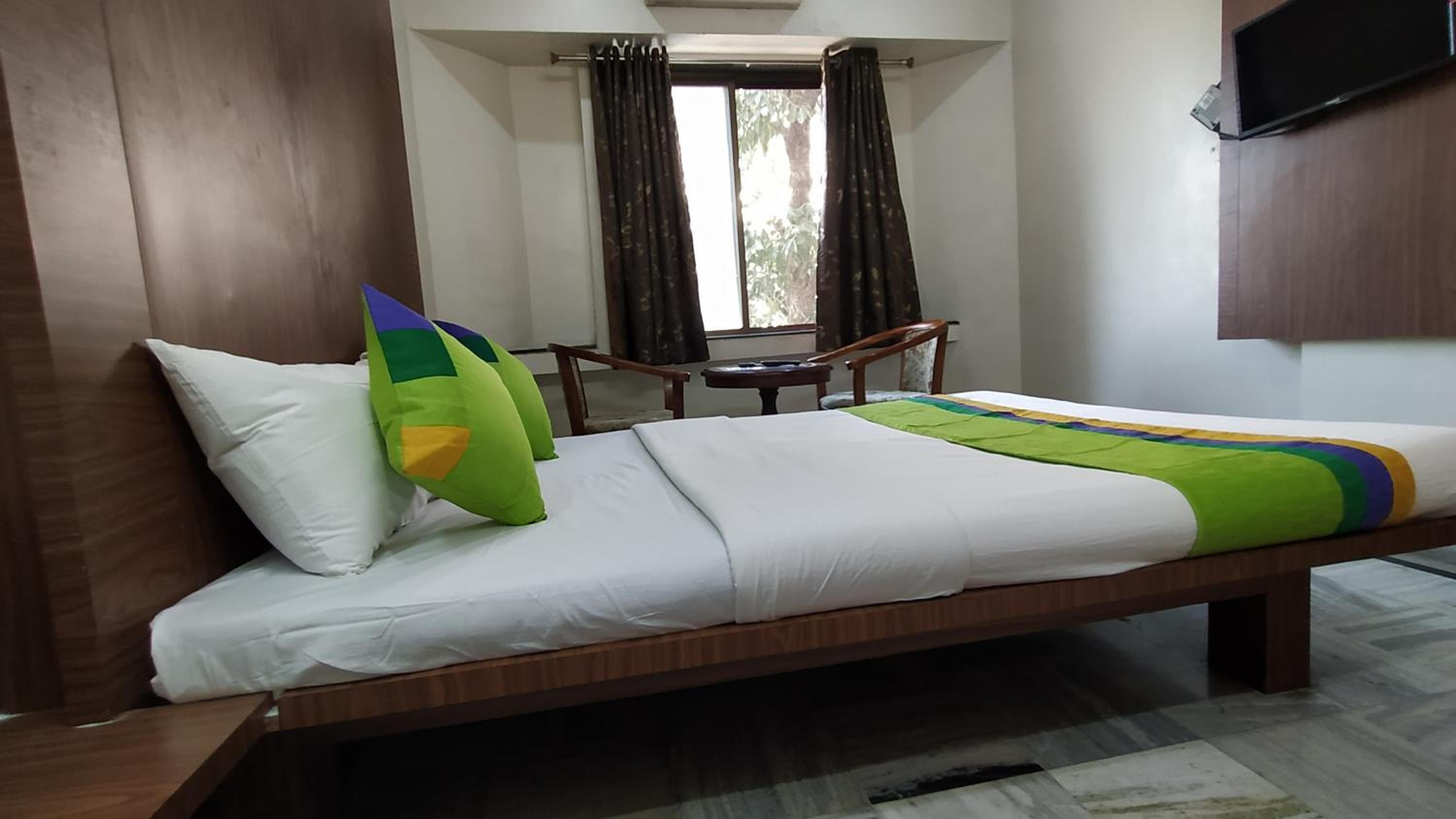 A hotel room with a wooden bed, white bedding, a green and yellow pillow, a small wooden table, and a TV mounted on the wall.