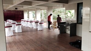 A room with rows of white chairs with red sashes, a wooden floor, a window with a view of trees, and a sink area. - Hotel City Palace (Nashik)